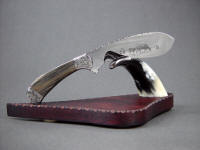 The Cattleman in etched 440C high chromium stainless steel blade, hand-engraved 304 stainless steel bolsters, Agatized Petrified Wood gemstone handle, on stand of polished cow horn, purpleheart (Amaranth) hardwood