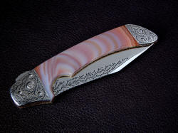 "Procyon" linerlock folding knife, obverse side view, folded. Knife has full engraving of light vine and tendril pattern on blade and bolsters. Handle scales are very hard and durable Brazilian Agate gemstone